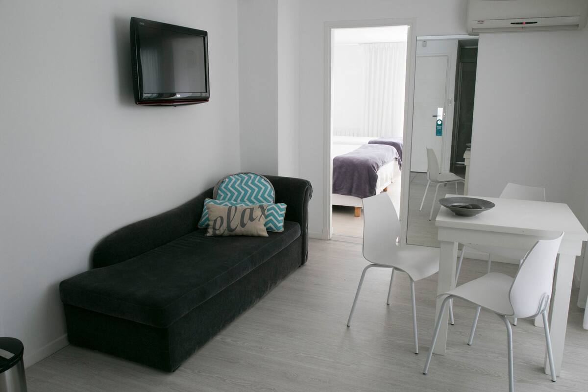 A modern, minimalistic room with a black sofa, white chairs, a small table, and a TV mounted on the wall.