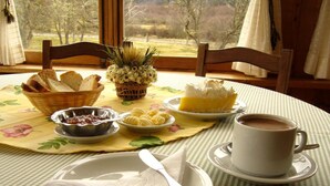 Desayuno con pan, bollería, mermelada, mantequilla y café sobre una mesa con vistas a árboles.