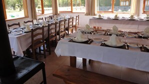 Comedor con mesas dispuestas para una comida, sillas de madera y ventanas grandes con cortinas.