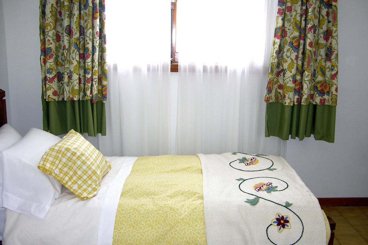 Ein Bett mit gelber Decke und Kissen, einem blumigen Überwurf und grünen Vorhängen.