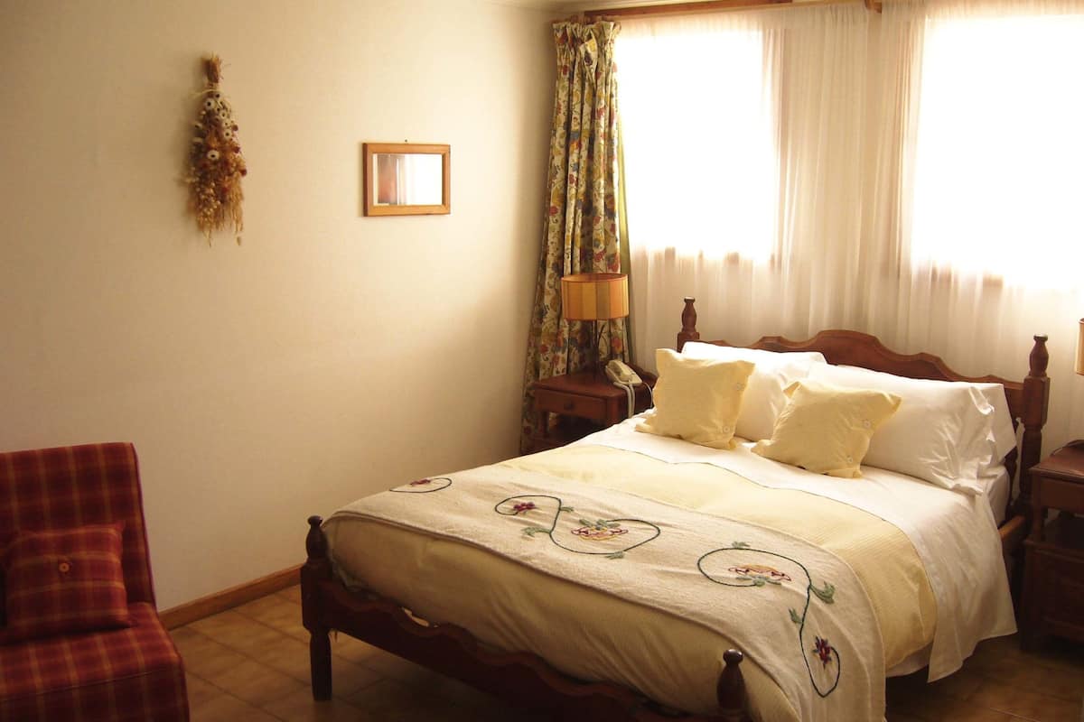 Ein Schlafzimmer mit Bett, Stuhl, Nachttisch, Lampe und einem Fenster mit Vorhängen.