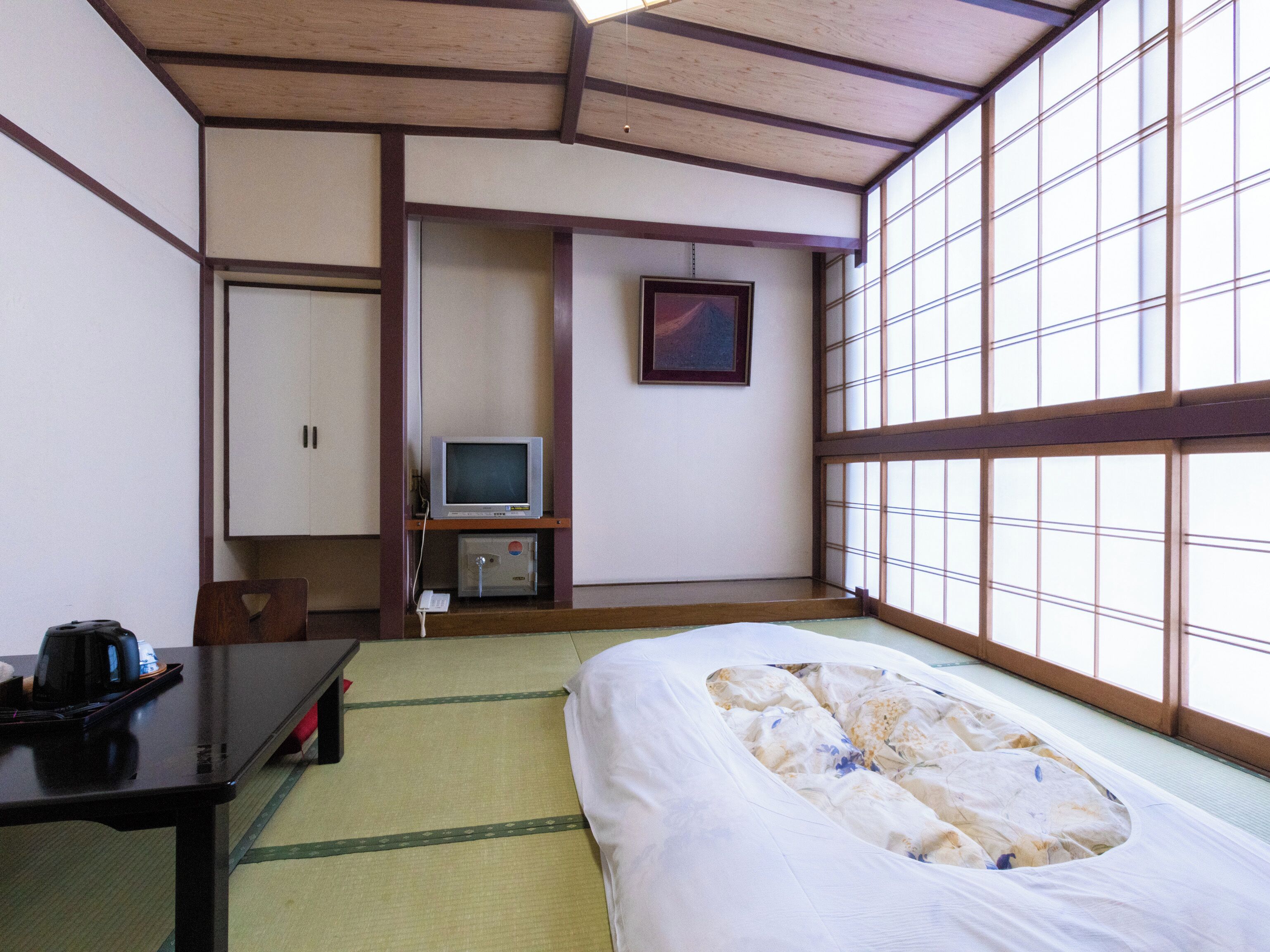 Quarto japonês tradicional com tatames, uma cama, uma escrivaninha com chaleira, televisão e um armário.