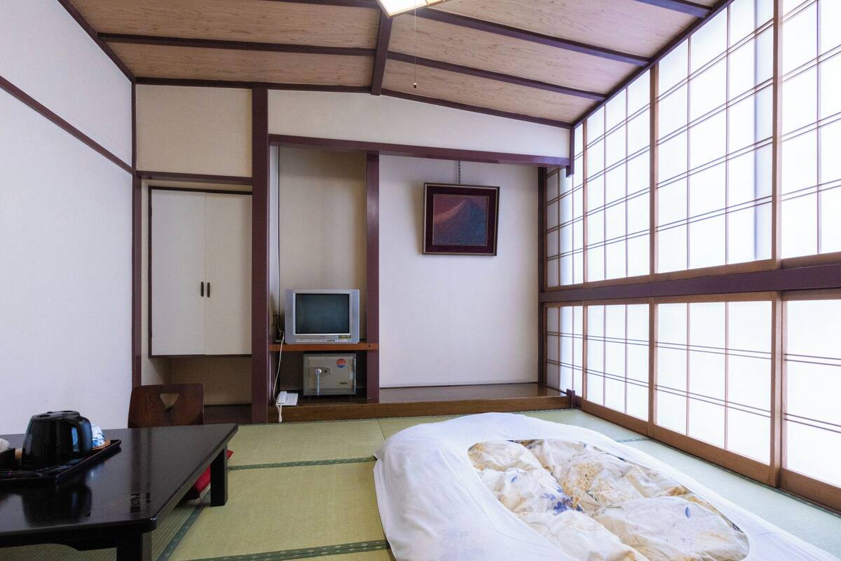 Ett traditionellt japanskt rum med tatamimattor på golvet, en säng, ett skrivbord med en vattenkokare, en tv och ett skåp.