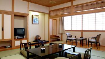 A traditional Japanese-style room with tatami flooring, a low table with chairs, a television, and a large window with shoji screens.