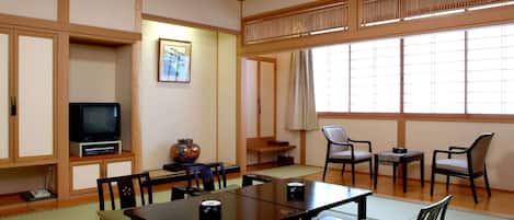 Une pièce de style japonais traditionnel, avec un sol recouvert de tatamis, une table basse entourée de chaises, un téléviseur et une grande fenêtre ornée de paravents shoji.