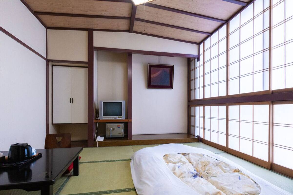 Ett traditionellt japanskt rum med tatamimattor på golvet, en säng, ett skrivbord med en vattenkokare, en tv och en skjutdörr.