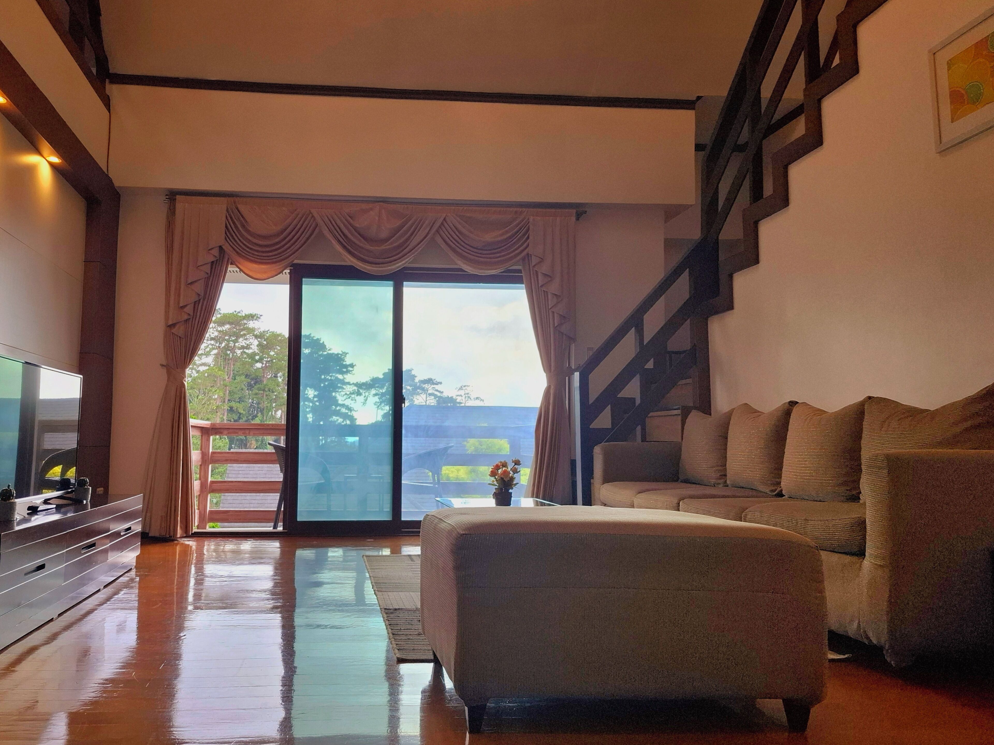 Un salón espacioso con un gran ventanal corredera que da a un balcón, un sofá cómodo, un televisor de pantalla plana y una escalera.