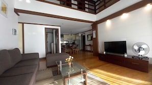 A living room with a sofa, a glass coffee table, a TV, and a fan. There is a kitchen in the background. - Prestige Vacation Apartments - Hanbi Mansions (Baguio)