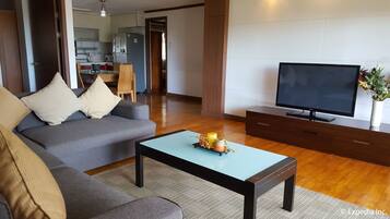 A modern living room with a grey sofa, a coffee table with decorative items, a flat-screen TV on a wooden stand, and a kitchen in the background.