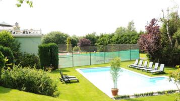 A swimming pool surrounded by a green lawn, a tennis court, and a fenced area with lounge chairs.