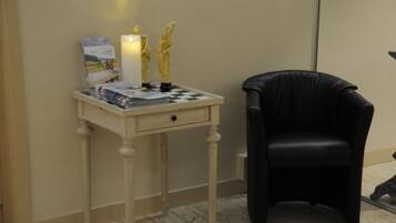 A small, neatly arranged room with a white bedside table, a black leather chair, a lit candle, and a chessboard.