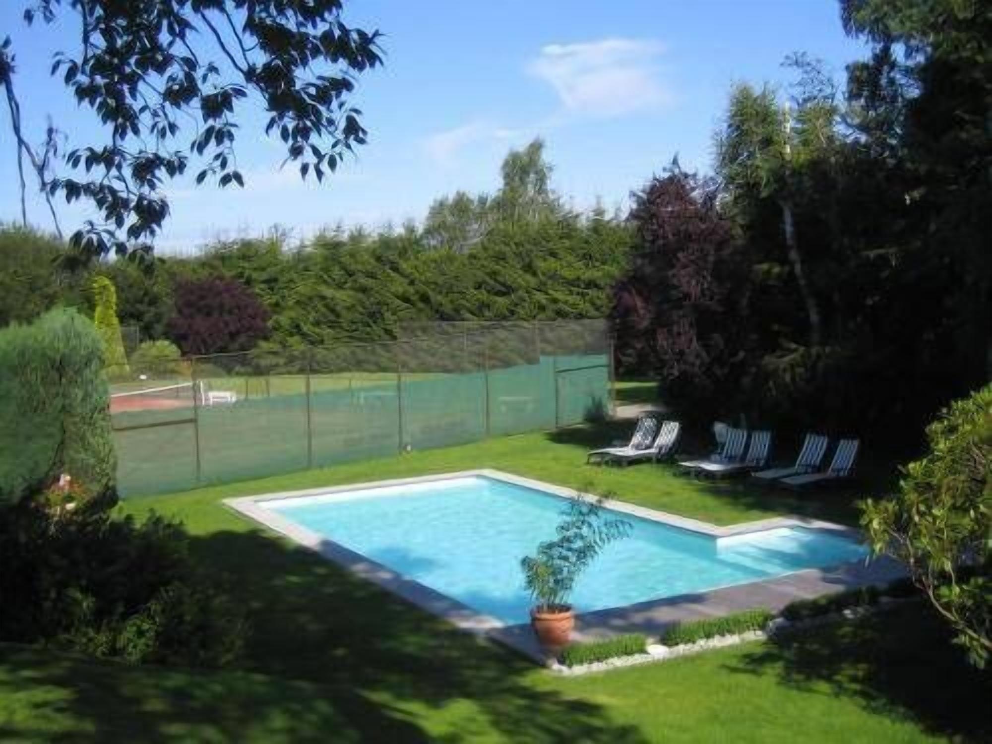 A rectangular swimming pool with a surrounding green lawn, a tennis court, and a row of lounge chairs.