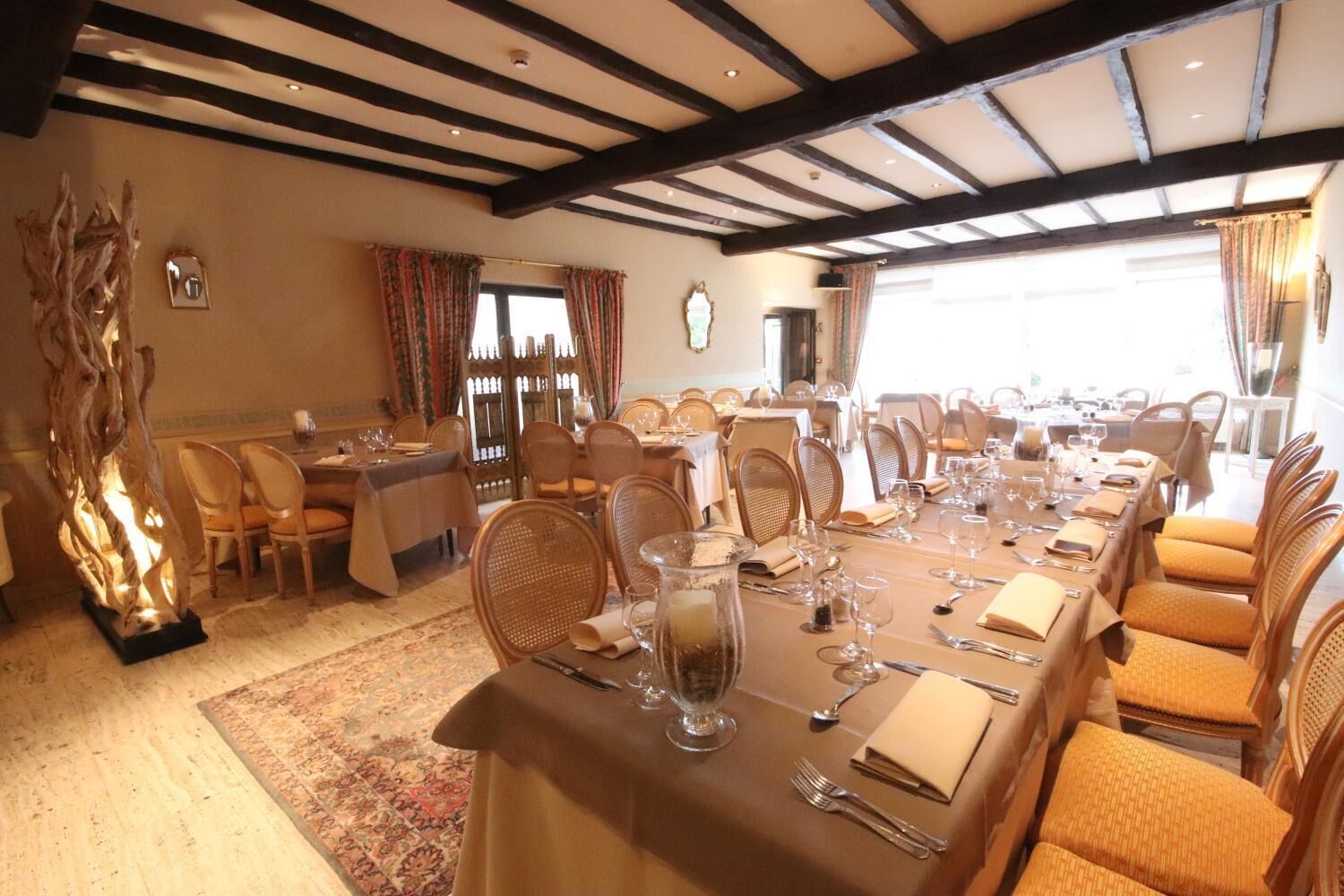 A dining area with multiple tables set for a meal, wooden beams on the ceiling, and a large window with curtains.