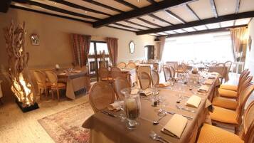 A dining area with multiple tables set for a meal, wooden beams on the ceiling, and a large window with curtains.