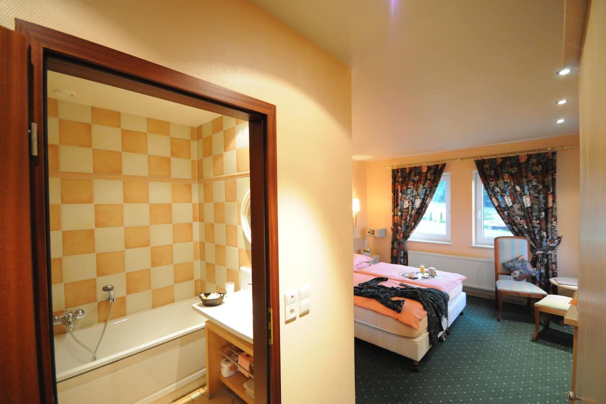 A hotel room with a checkered shower wall, a bed with a pink blanket, and a chair near a window with curtains.