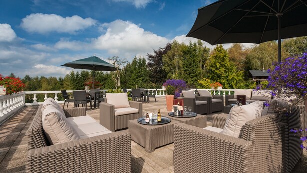 An outdoor patio with wicker furniture, umbrellas, and a view of trees and flowers.