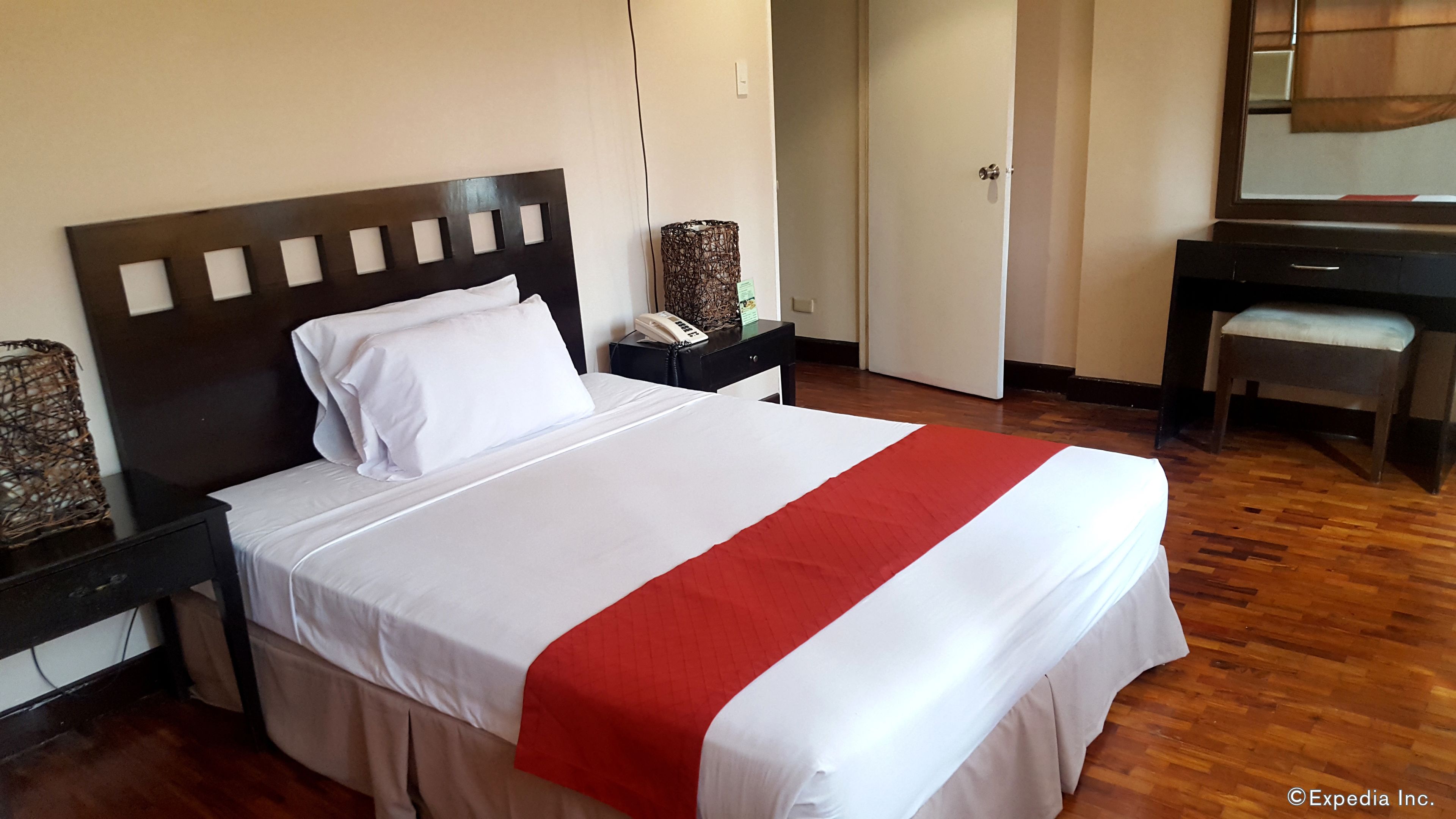A neatly made bed with a white and red bedspread, a wooden headboard, a bedside table with a telephone, and a desk with a chair.