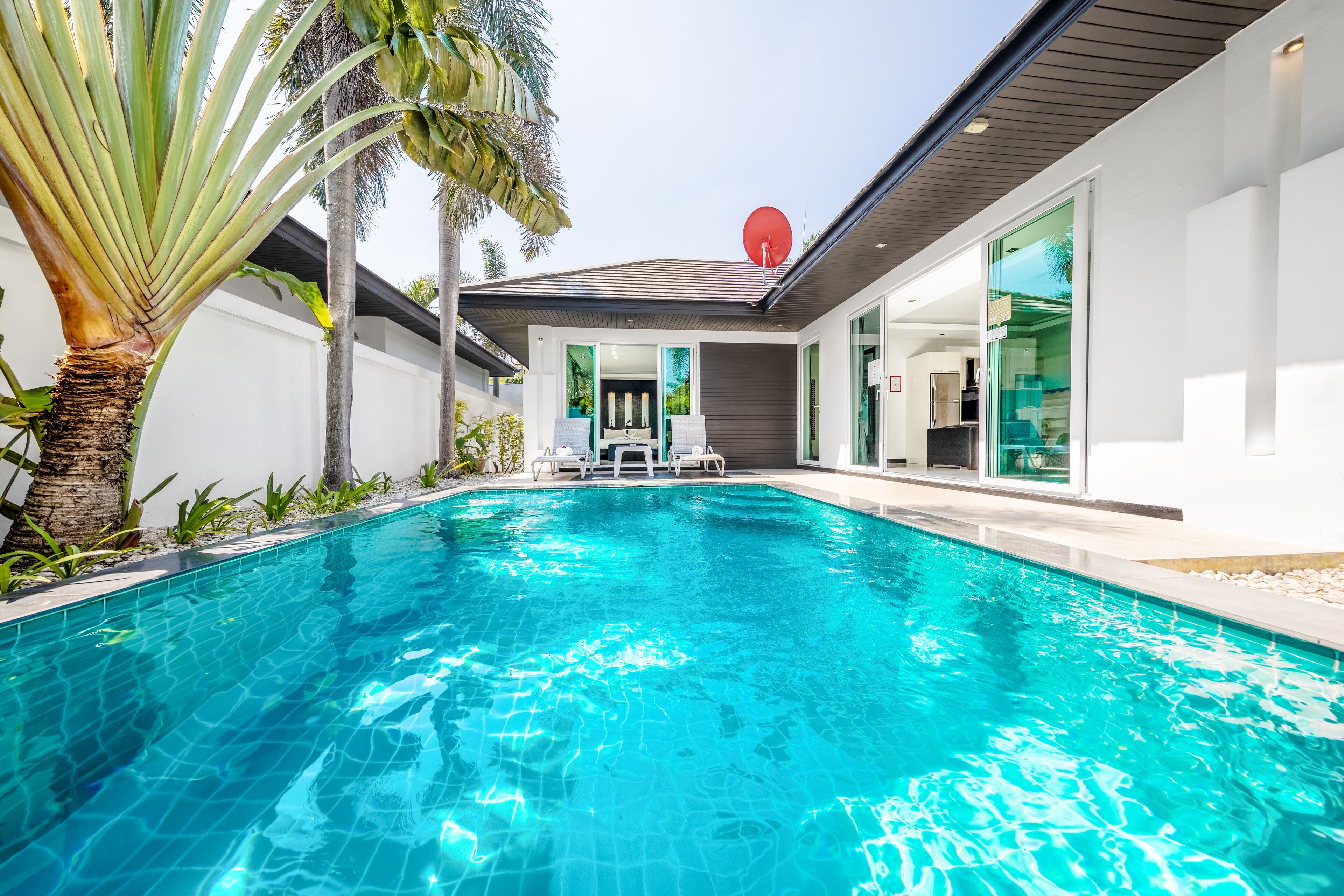 A swimming pool with clear blue water, surrounded by a modern house with large windows and a satellite dish.
