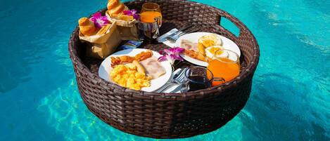 A wicker basket filled with breakfast items including eggs, bacon, toast, and juice, placed on a poolside.