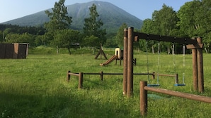 Un terrain de jeux avec des balançoires et un toboggan, situé devant un paysage de montagne et de forêts.