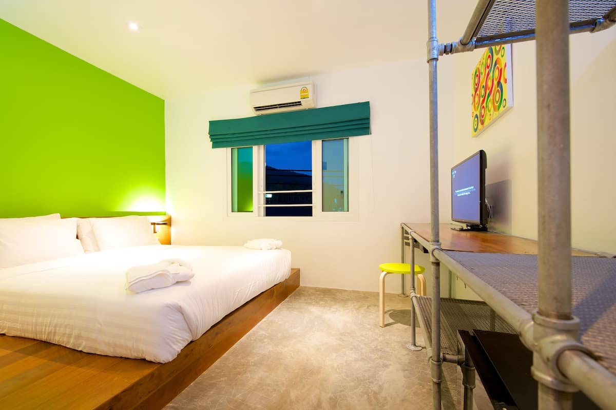 A hotel room with a bed, a desk with a computer, an air conditioner, and a window with a view of the outside.