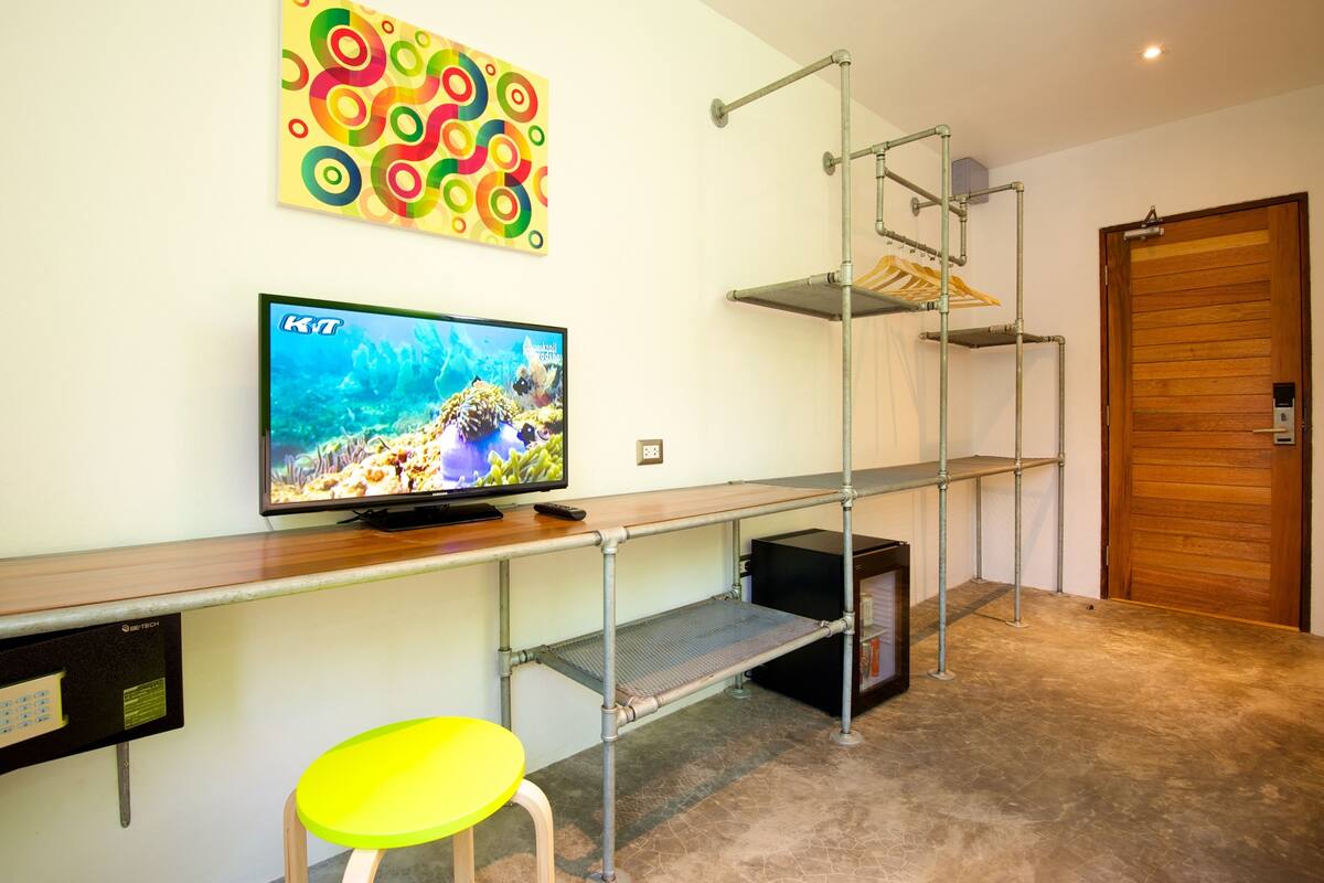 A room with a TV on a wooden shelf, a metal shelf unit, a yellow chair, and a wooden door.
