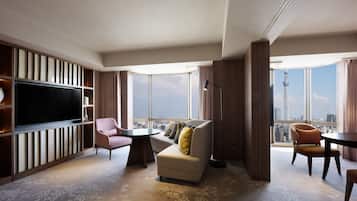 A modern hotel room with a large flat-screen TV, a sofa, a chair, and a small table. The room has a large window with a city view.