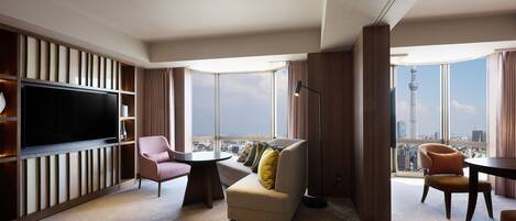 Habitación de hotel moderna con una televisión de pantalla plana grande, un sofá, una silla y una mesita pequeña. La habitación tiene un ventanal con vistas a la ciudad.