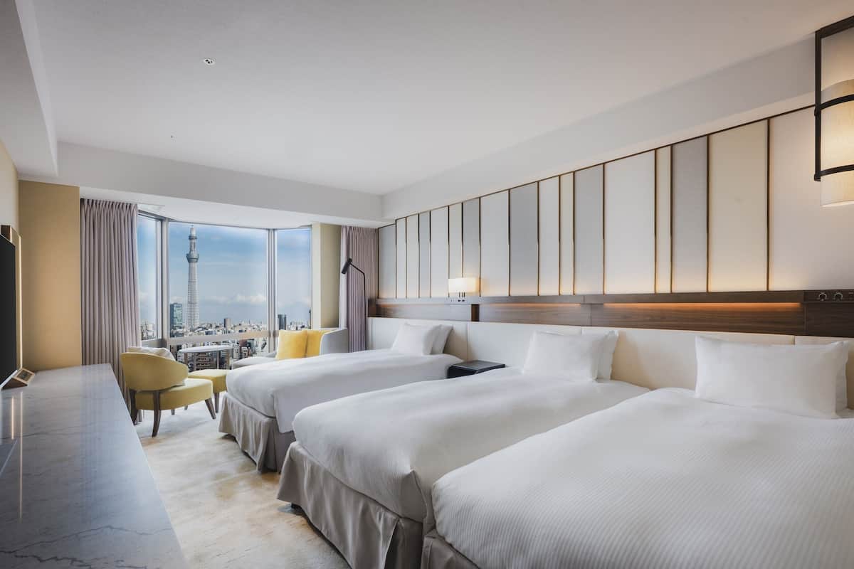 A hotel room with two beds, a desk, a chair, and a large window with a city view.