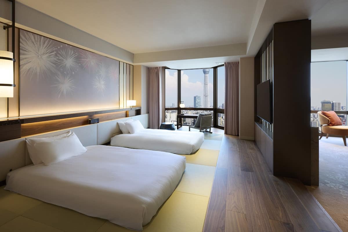 A hotel room with two beds, a large window with a city view, a desk with a chair, and a TV.