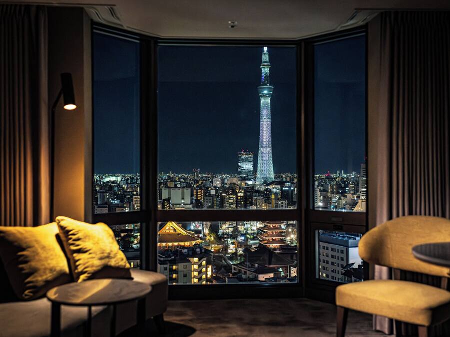 A corner room with a large window overlooking a cityscape at night, featuring a prominent tower.