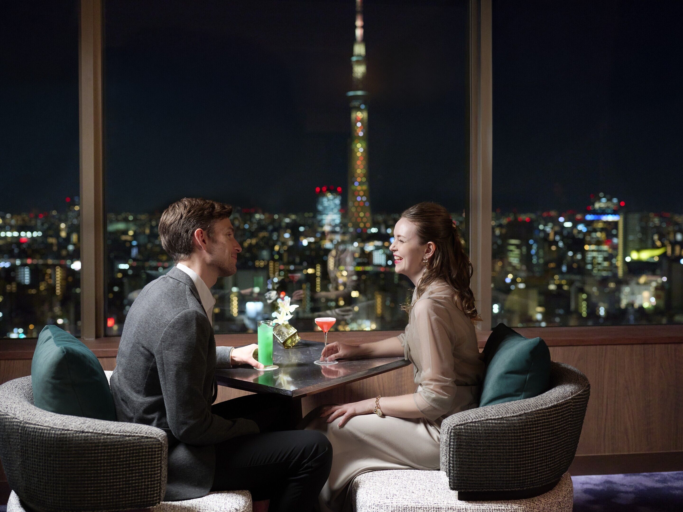 Um casal sentado à mesa com bebidas, com vista para um horizonte urbano e uma torre imponente.