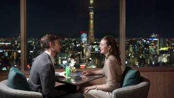 Um casal sentado à mesa com bebidas, com vista para um horizonte urbano e uma torre imponente.