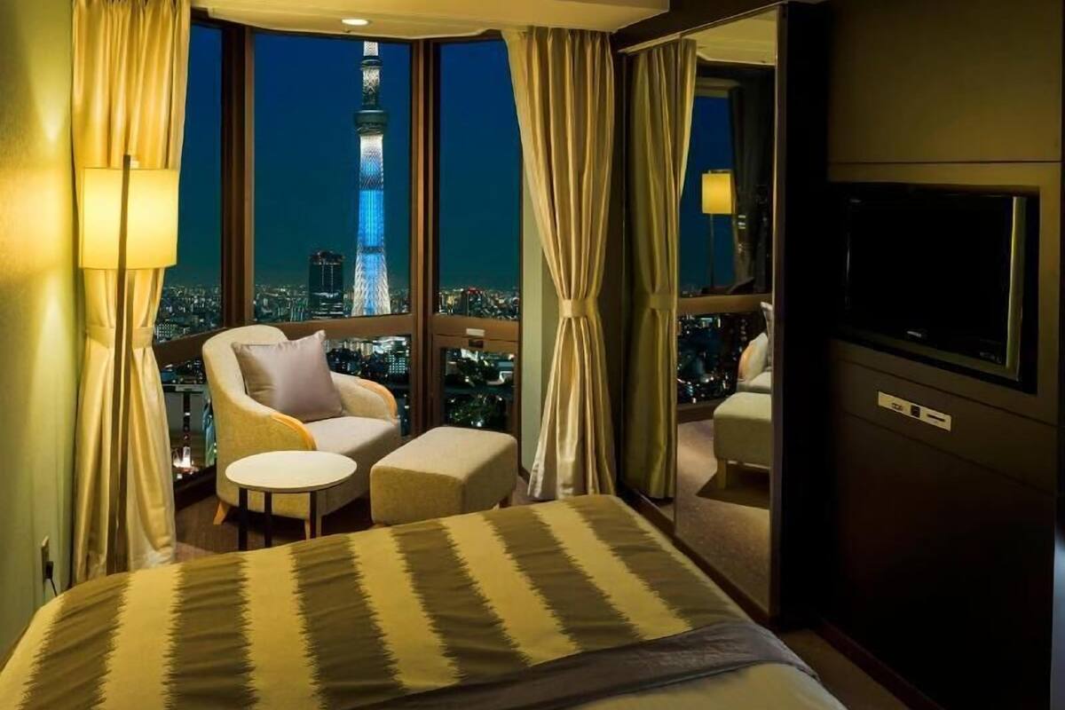 A hotel room with a bed, a sofa, a small table, a TV, and a view of a cityscape at night.