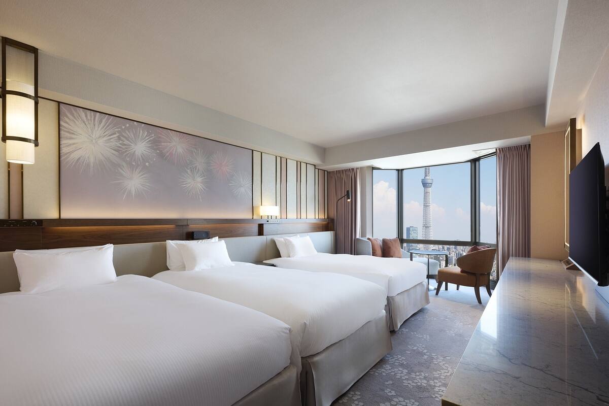 A hotel room with two beds, a desk, and a large window offering a city view.