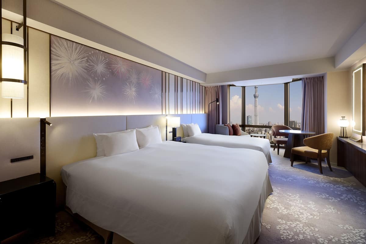 A hotel room with two large beds, a desk, a chair, and a view of a cityscape including a tower.