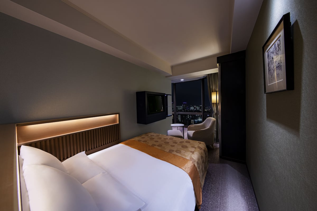 A hotel room with a large bed, a desk, a chair, a TV, and a city view.