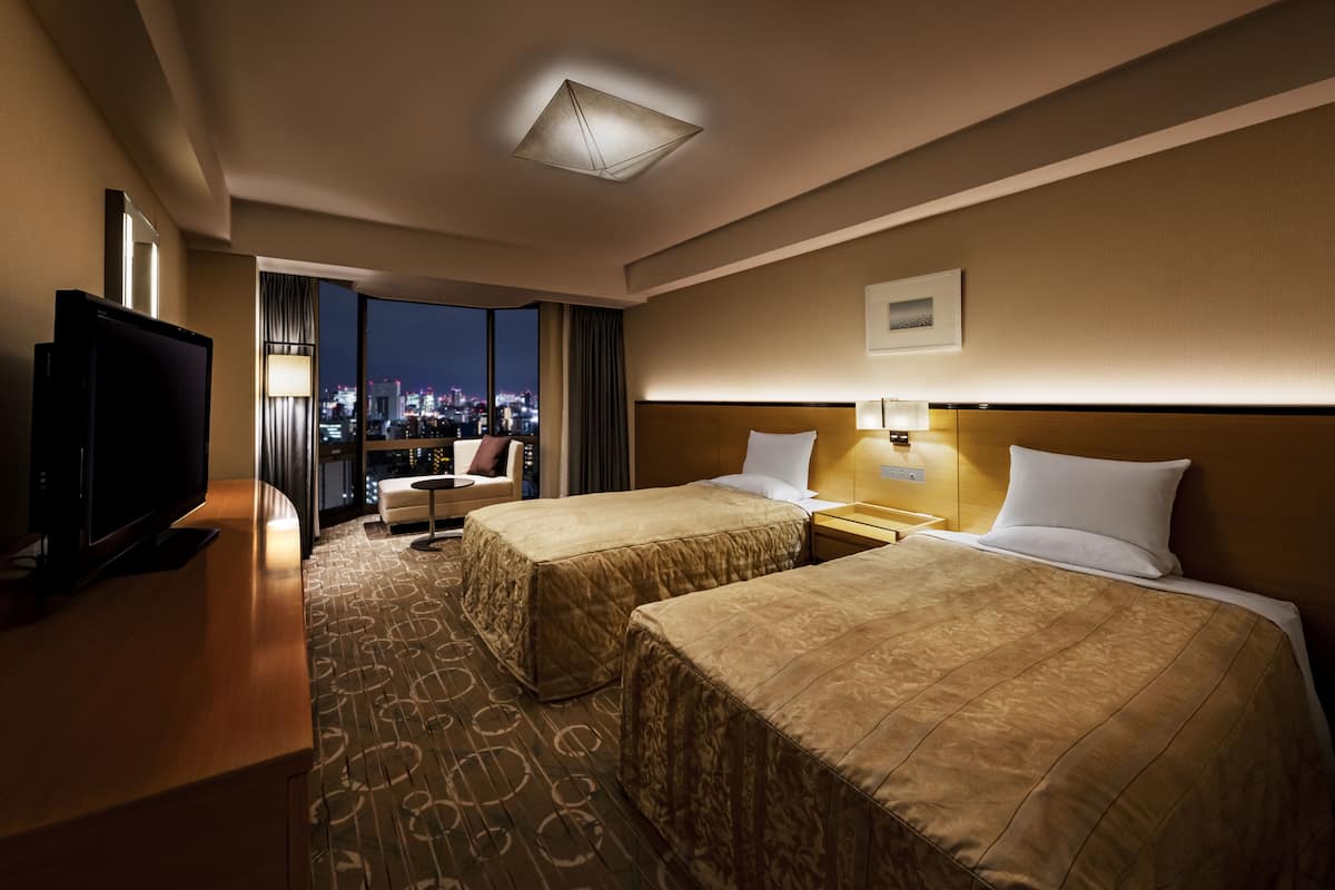 A hotel room with two beds, a TV, a desk, and a view of the city at night.