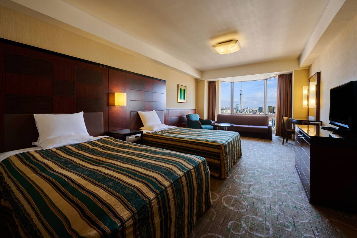 A hotel room with two beds, a desk, a chair, a TV, and a large window offering a city view.