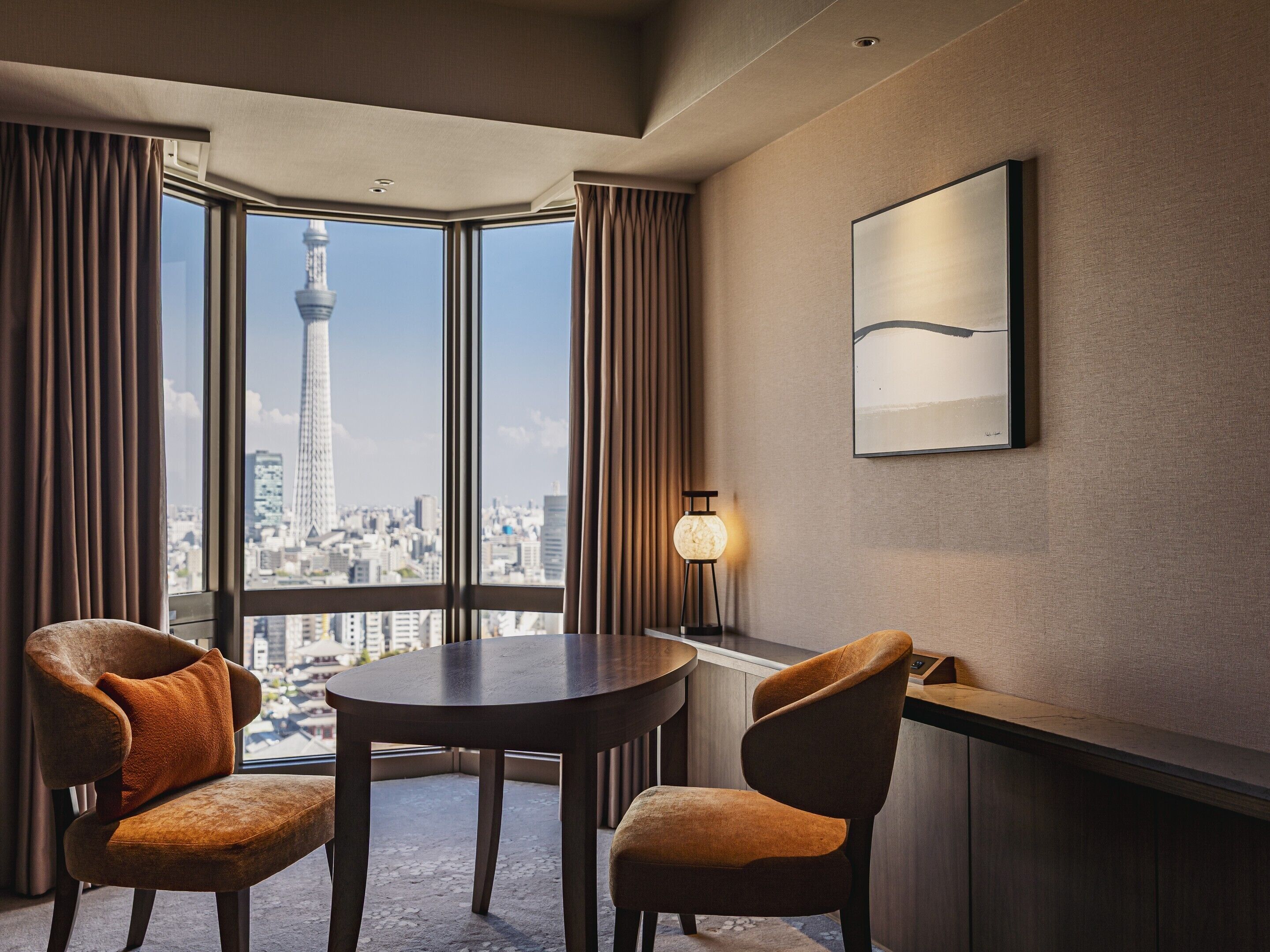 A modern hotel room with a large window offering a city view, a round table, two chairs, a floor lamp, and a framed artwork on the wall.