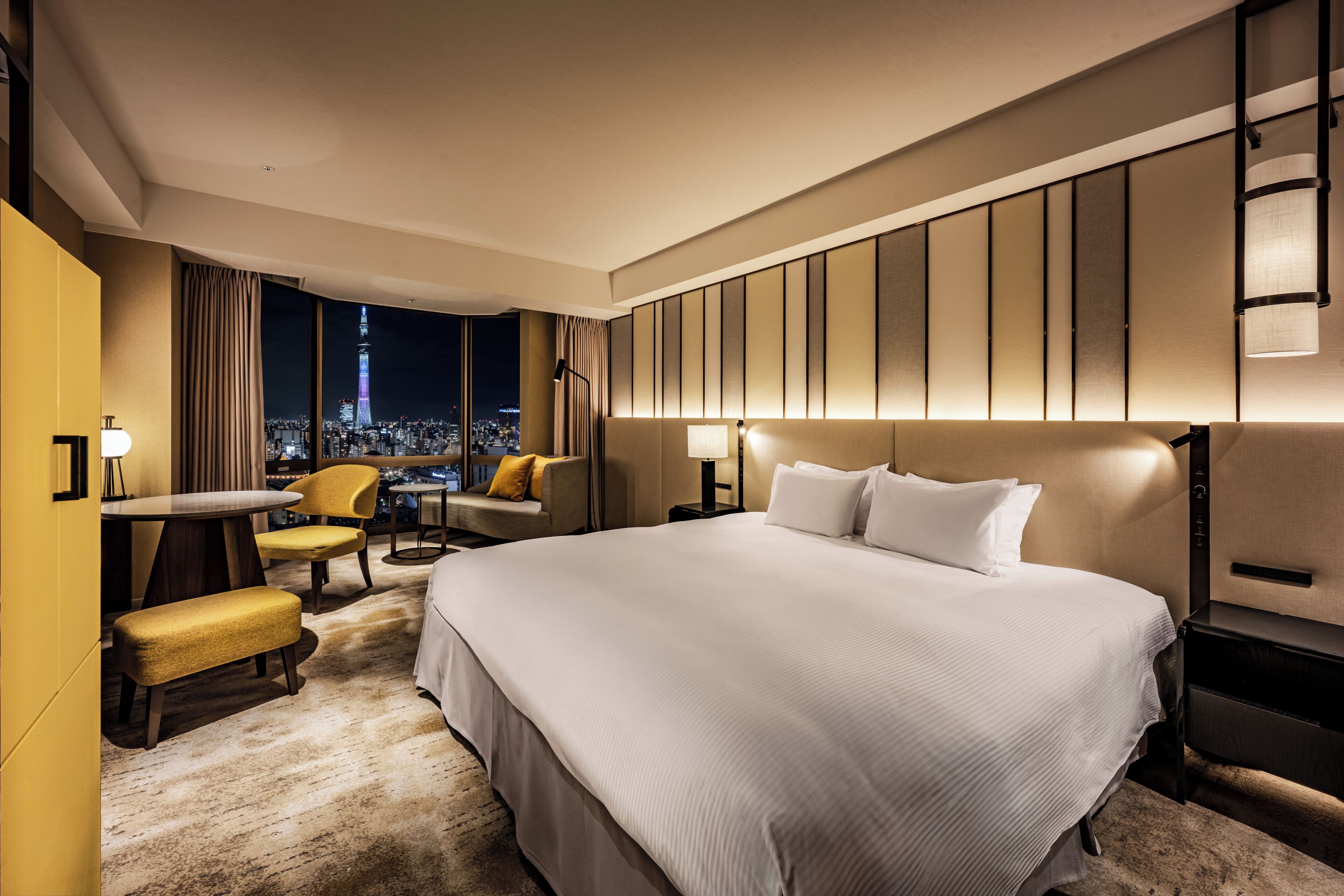 A hotel room with a large bed, a desk, two chairs, a bench, and a view of a cityscape at night.