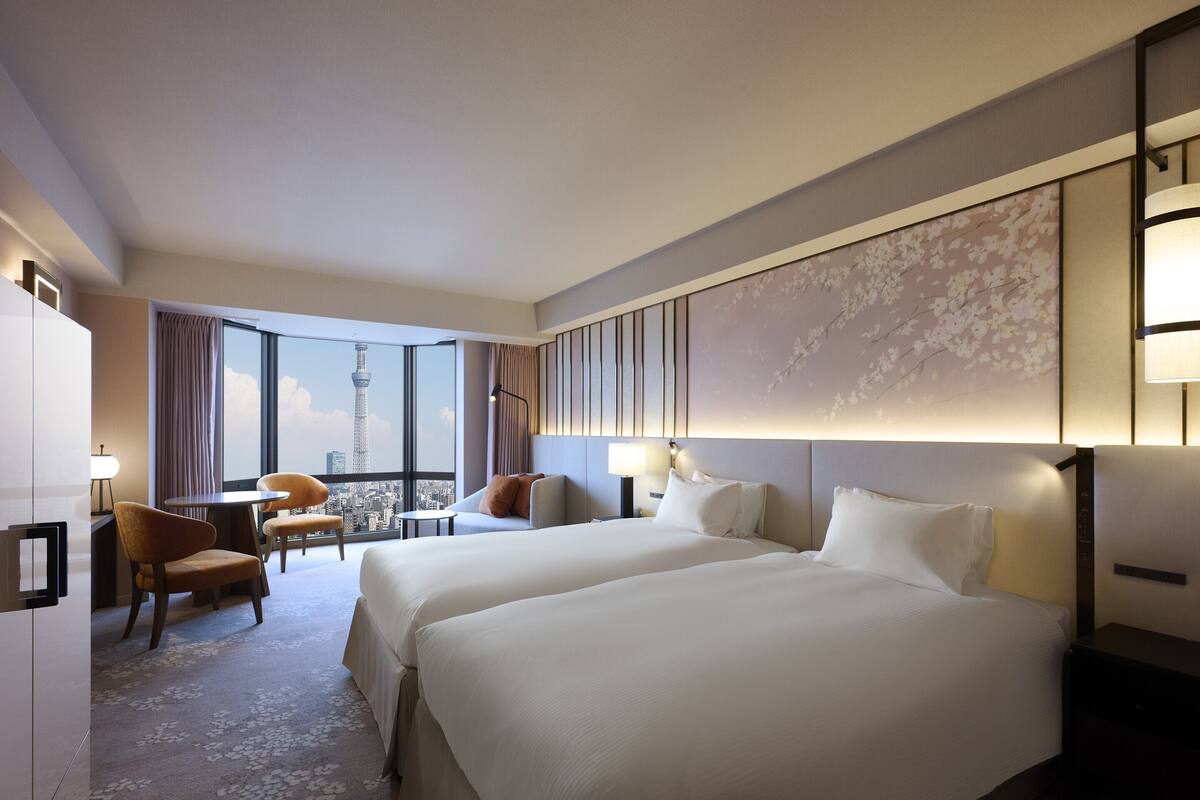 A hotel room with two beds, a desk, a chair, and a view of a cityscape.