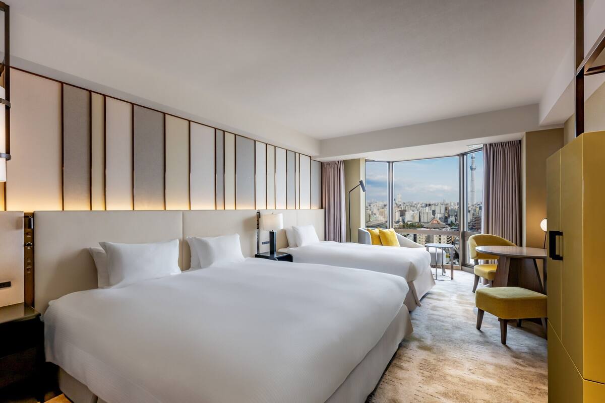 A hotel room with two beds, a desk, a chair, a view of the city, and a large window.