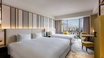 A hotel room with two beds, a desk, a chair, a view of the city, and a large window.
