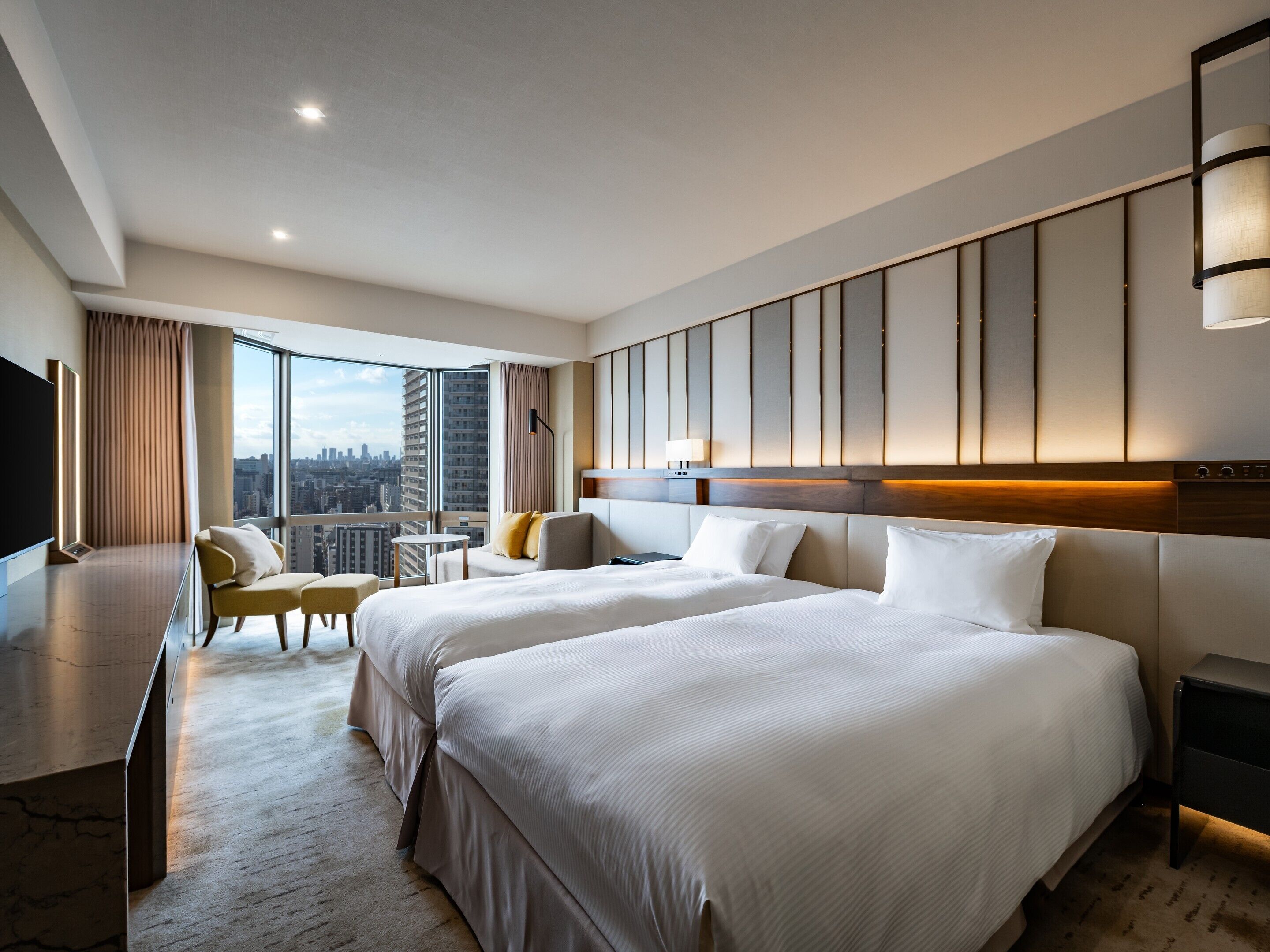 A modern hotel room with two large beds, a seating area, a TV, and a city view.