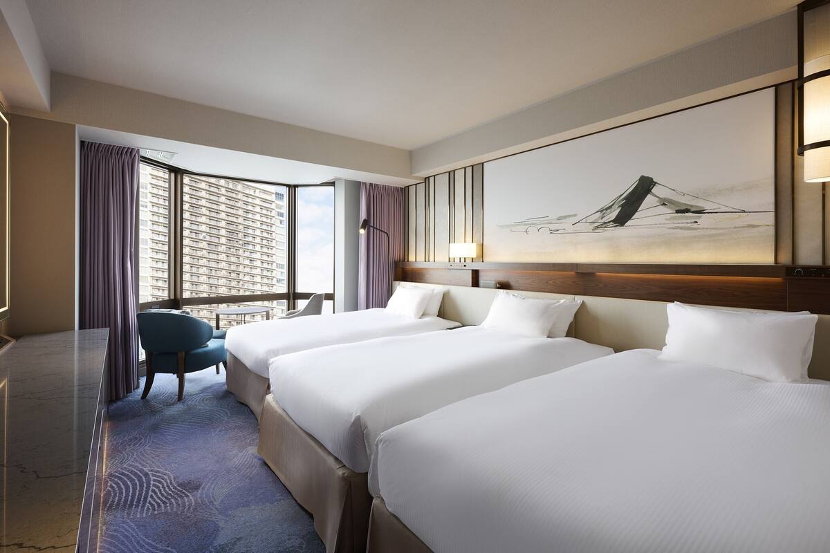 A hotel room with two beds, a desk, a chair, and a large window with curtains.