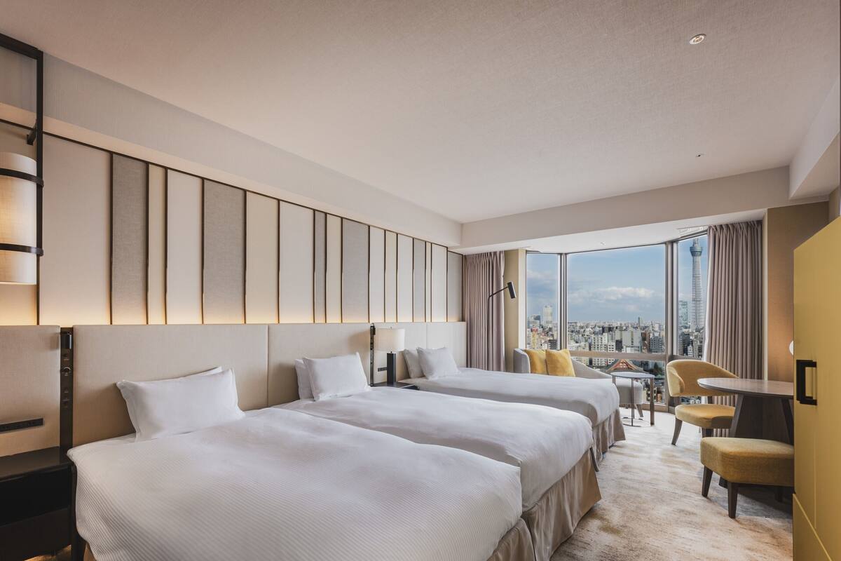A hotel room with two beds, a desk, a chair, and a large window with a city view.