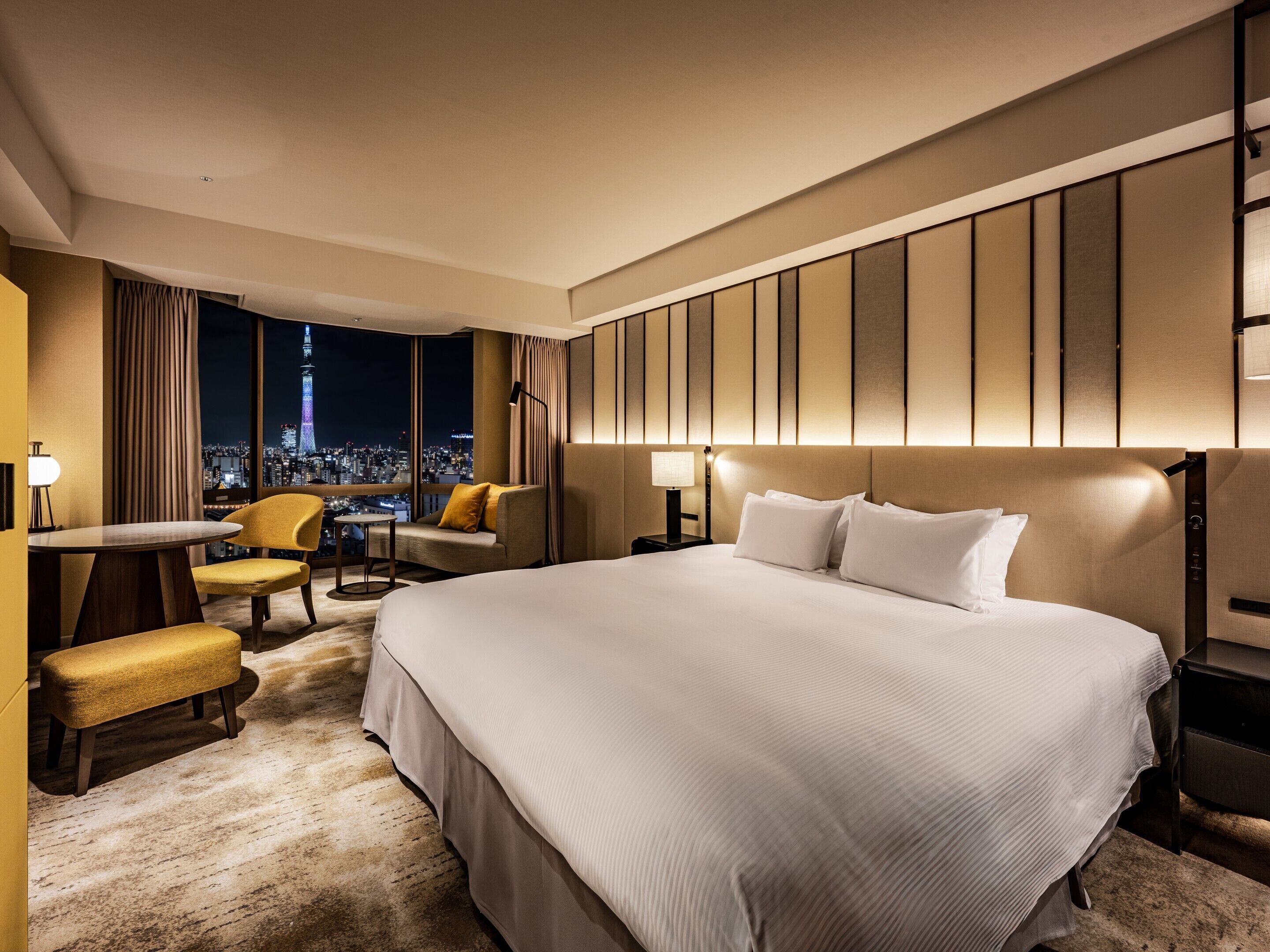 A hotel room with a large bed, a desk with a chair, a sofa, and a view of a cityscape at night.
