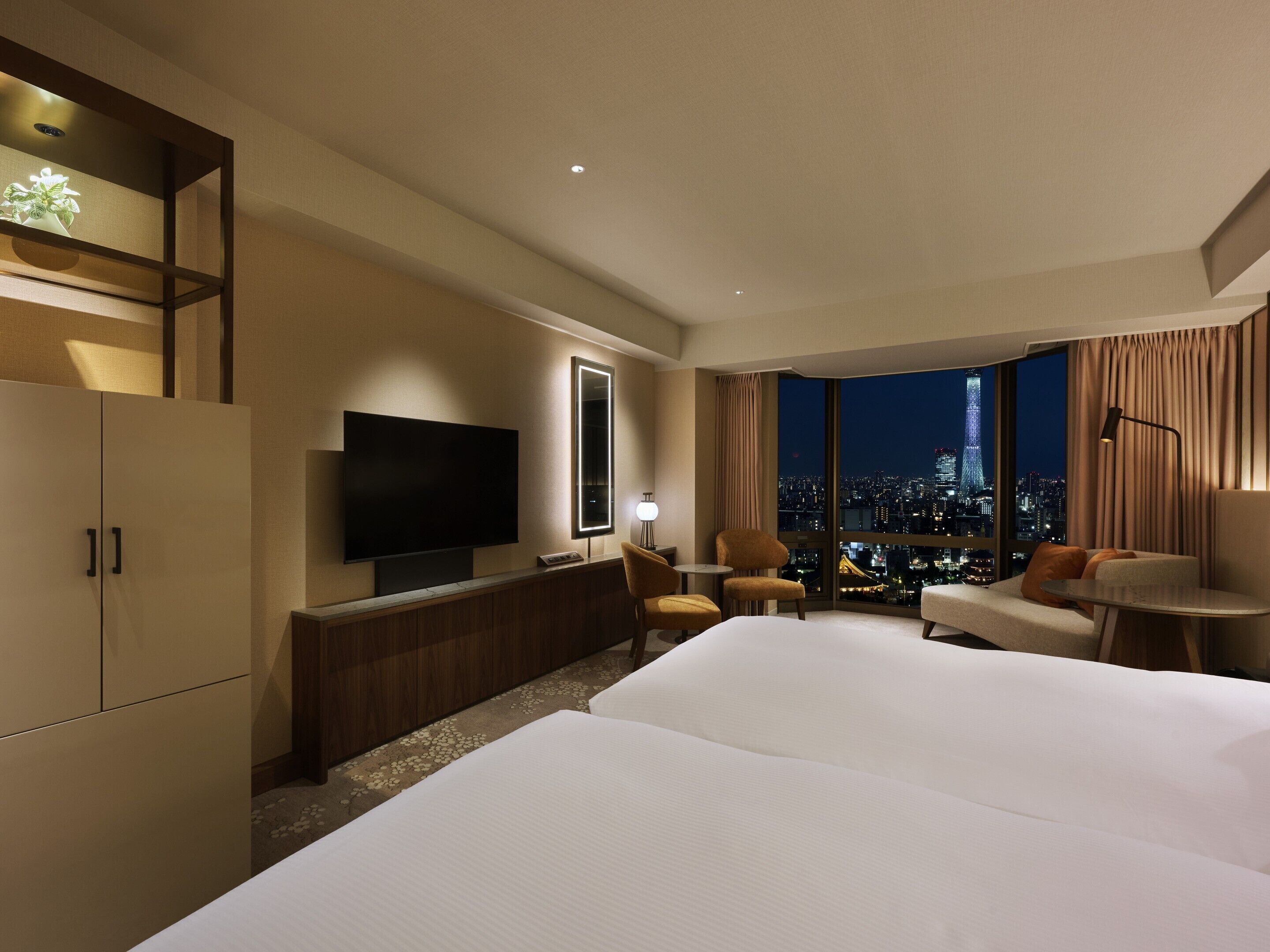 A hotel room with a large bed, a TV, a desk, and a chair. There is a view of a cityscape through the window.
