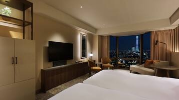 A hotel room with a large bed, a TV, a desk, and a chair. There is a view of a cityscape through the window.